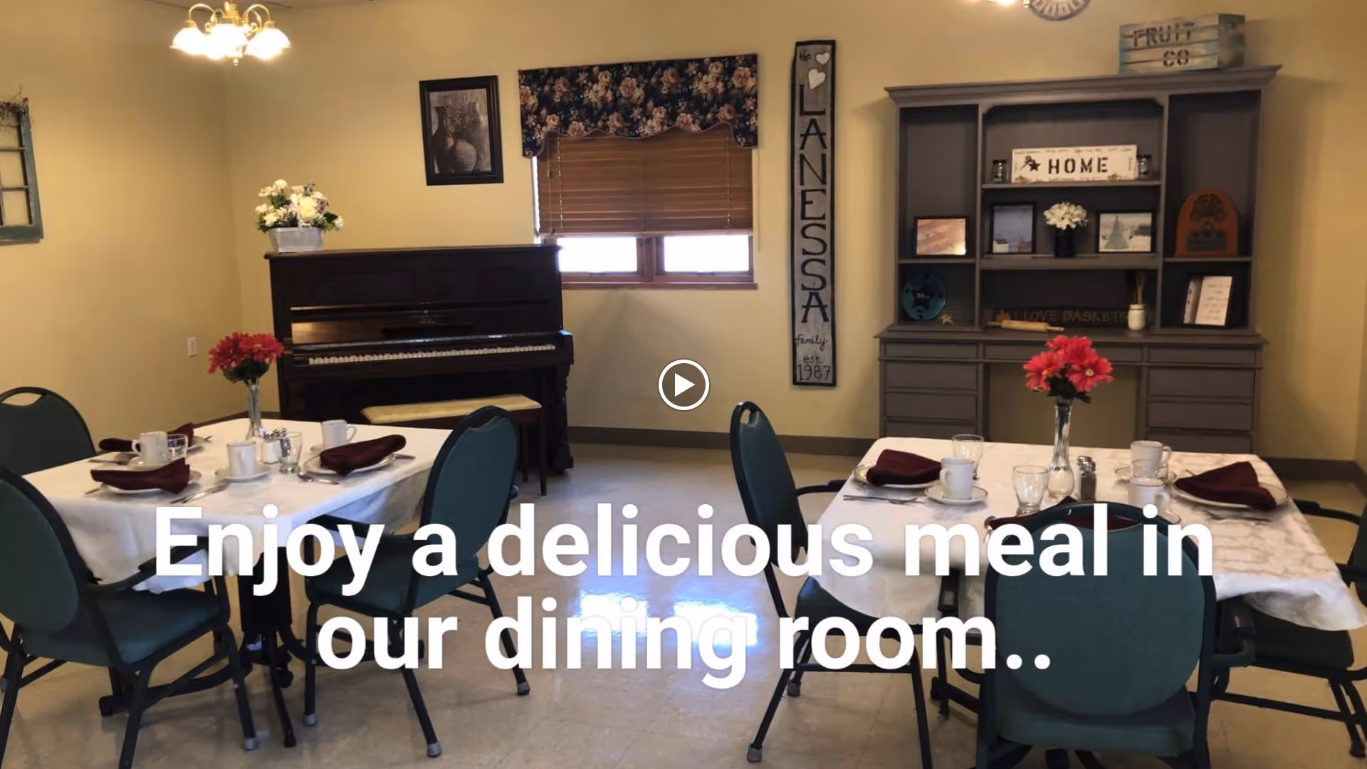 A dining room with two tables set for meals, each with four green chairs. The tables have white tablecloths, cups, glasses, and napkins. In the background, there is a piano, a window with a floral valance, and a gray hutch with decorative items. A vertical sign on the wall reads 'LANESSA Family est. 1987'.