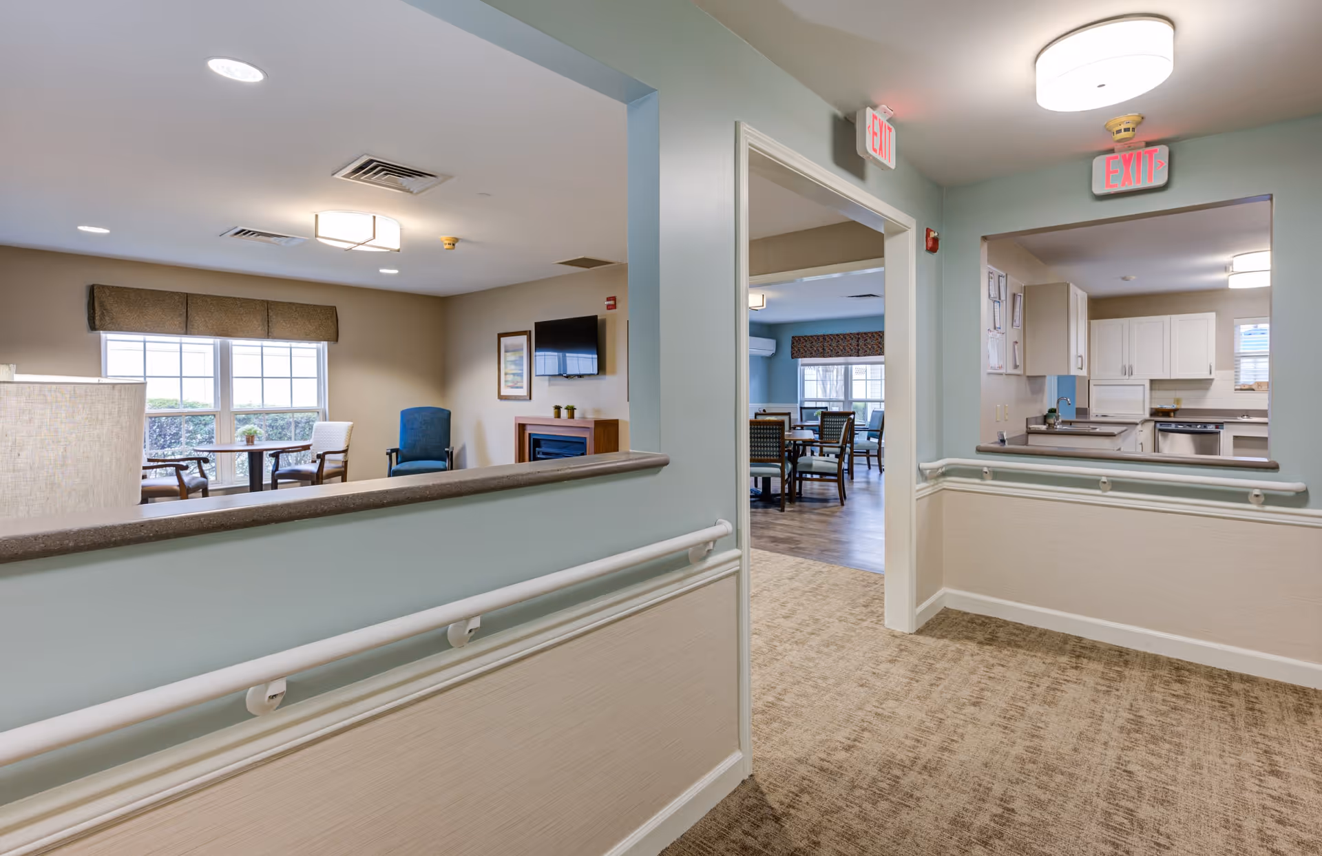 Interior view of a senior living facility hallway with handrails on the walls. The hallway opens into a living room area with chairs, a table, a TV mounted above a fireplace, and large windows with valances. Another opening shows a kitchen area with white cabinets and appliances. The space is well-lit with ceiling lights and has exit signs above the doorways.