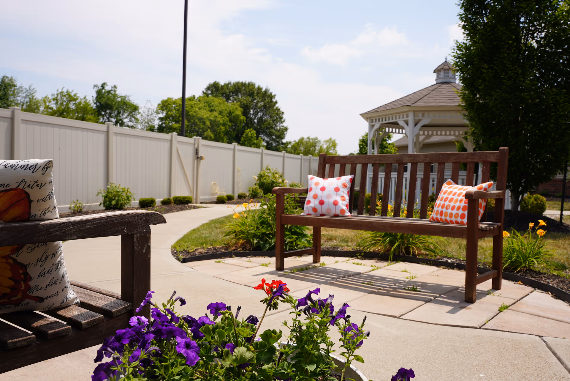 Outdoor patio with wooden benches, colorful pillows, flowering plants, a paved walkway and a gazebo in the background.
