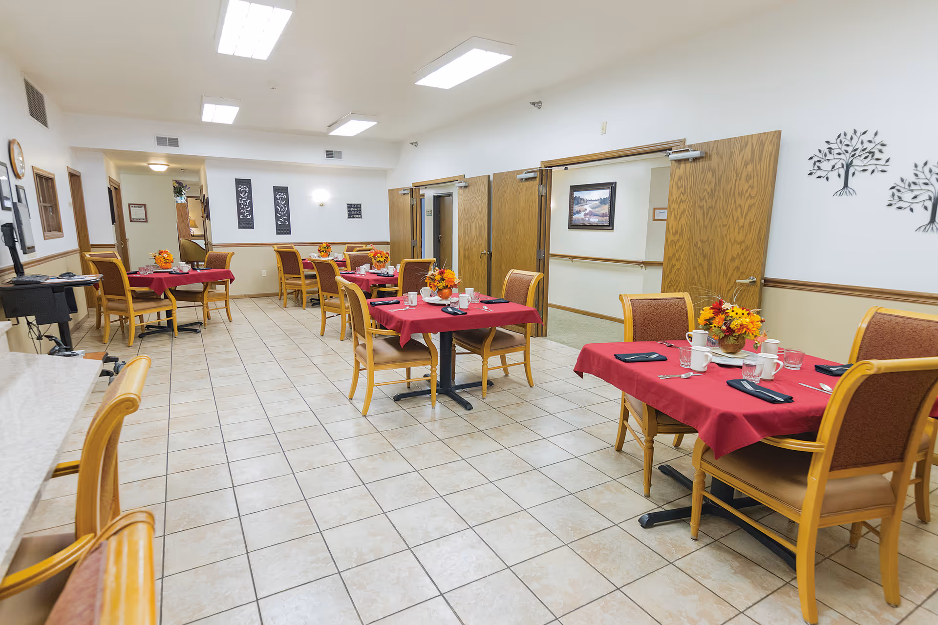 Dining room with multiple tables set with red tablecloths, chairs, and floral centerpieces in a senior living facility.