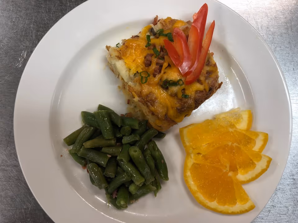 A white plate with a serving of cheesy casserole topped with chopped green onions and red bell pepper slices, a side of cooked green beans, and several slices of fresh orange.