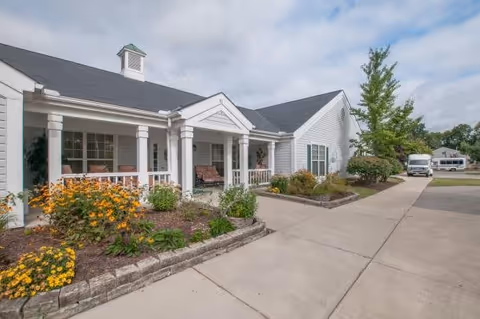 Exterior view of a single-story white building with a covered porch featuring seating. There are flower beds with yellow and orange flowers along the front, a concrete walkway, and a tree on the right side. The sky is partly cloudy.