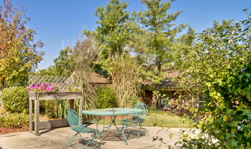 Outdoor patio area with a green metal table and two matching chairs surrounded by lush greenery, trees, and flowering plants under a clear blue sky.