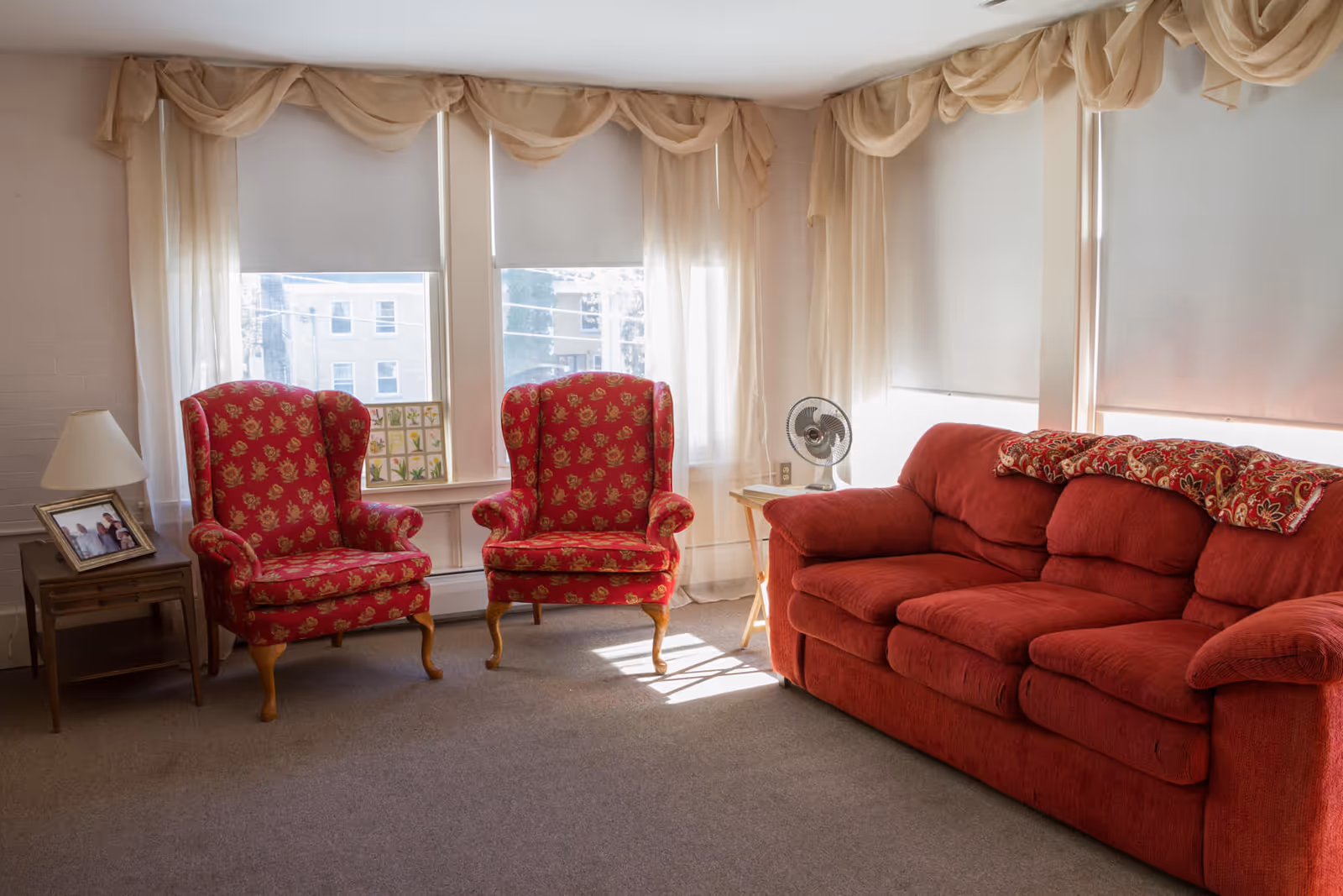 A cozy living room with two red patterned armchairs and a red sofa. The room has large windows with sheer curtains and roller blinds, a small side table with a lamp and a framed photo, and another small table with a fan. Sunlight is streaming into the room.