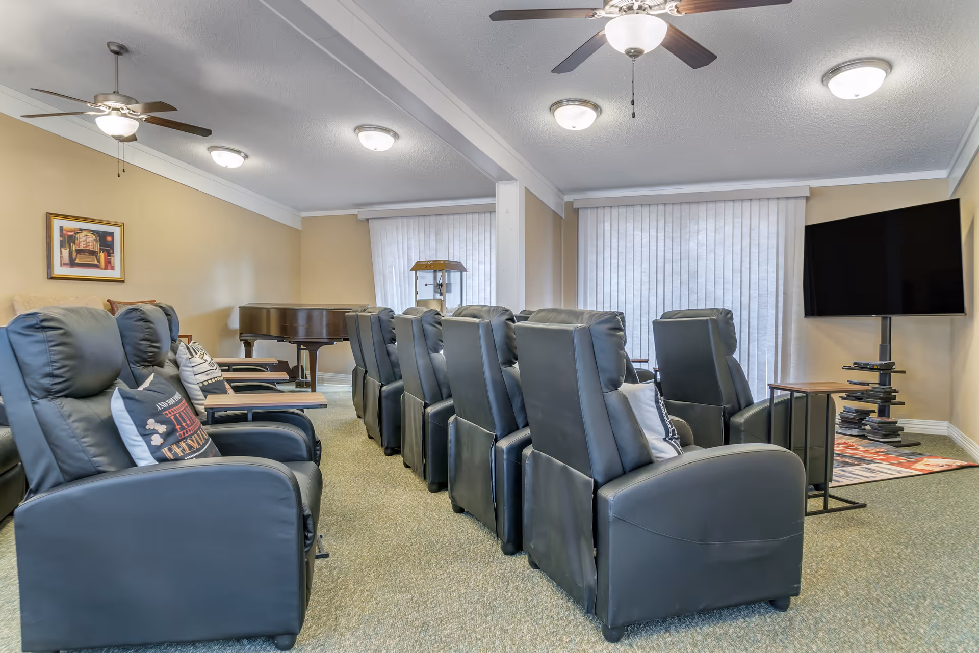 A row of black reclining chairs in a common lounge facing a large wall-mounted TV with a piano in the background.