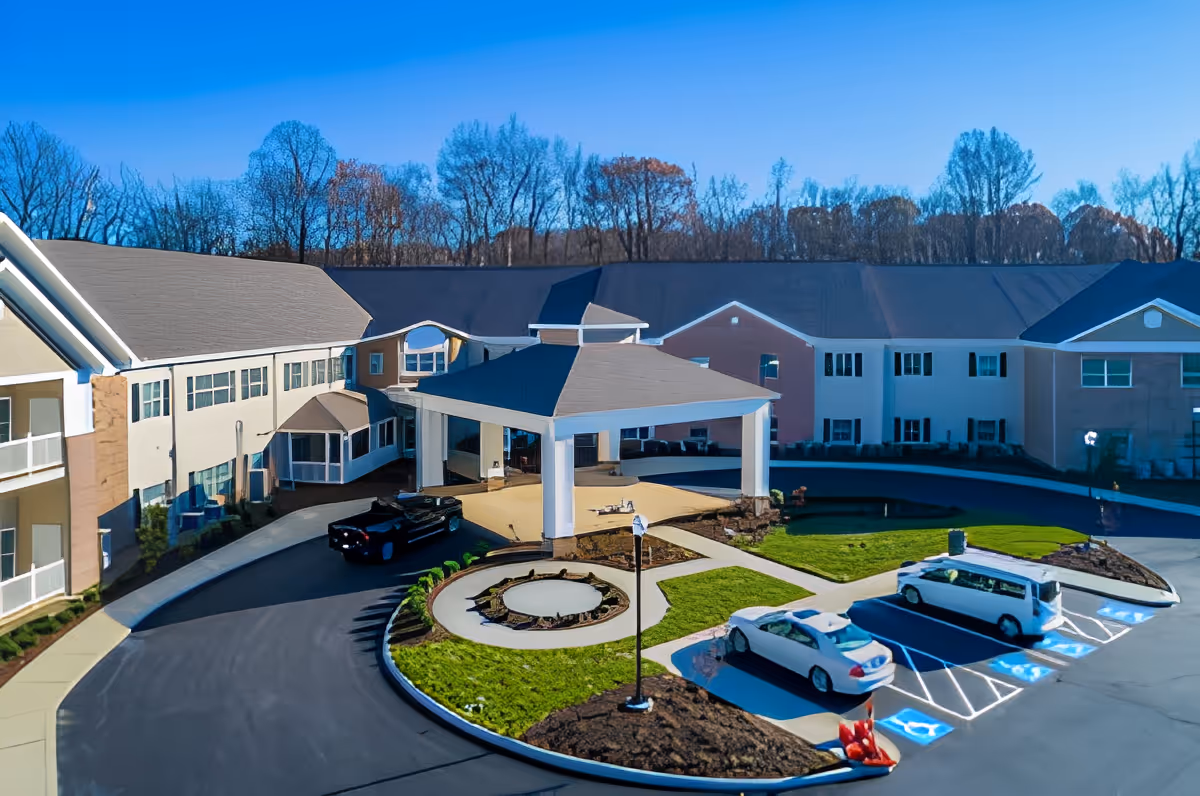 Exterior view of Danbury Brunswick senior living facility showing a large two-story building with a covered entrance driveway, several parked cars including a white van, landscaped greenery, and trees in the background under a clear blue sky.
