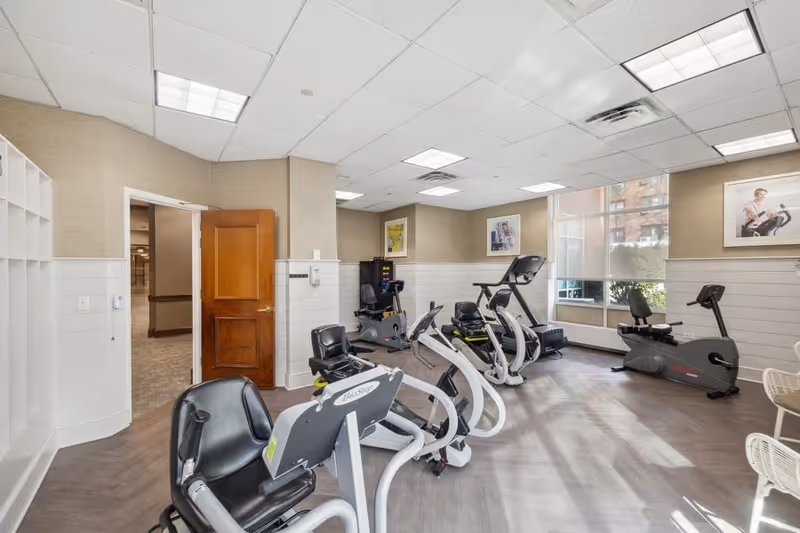A well-lit exercise room with several stationary exercise bikes and a treadmill. The room has large windows letting in natural light, beige walls with white wainscoting, and framed pictures on the walls. There is a wooden door open to an adjacent hallway.