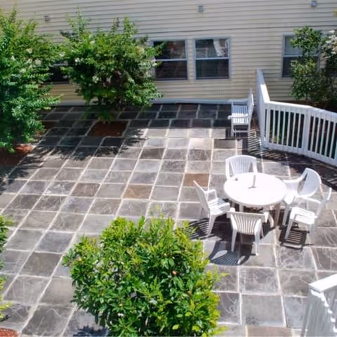 Outdoor patio area with a tiled floor, a round white table surrounded by five white chairs, some green bushes, and a building wall with windows in the background.