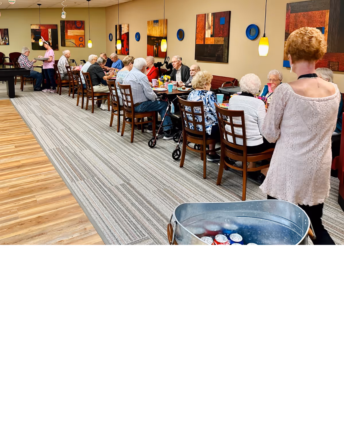 A group of seniors seated at a long communal dining table with staff serving and artwork on the walls.
