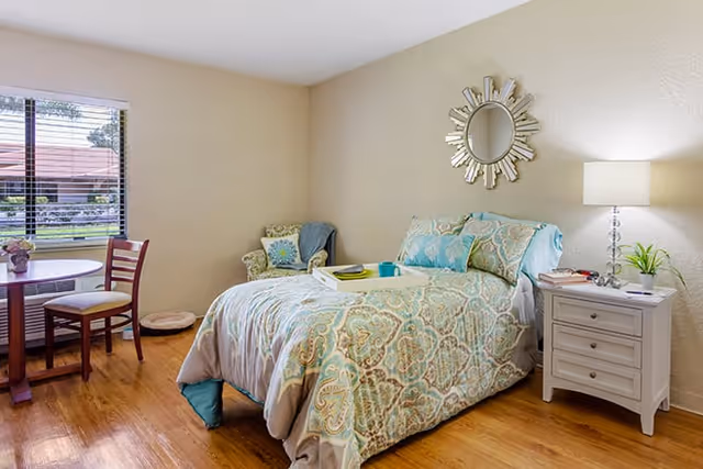 A cozy bedroom with a bed covered in a patterned comforter and pillows, a white nightstand with a lamp and plant, a decorative round mirror on the wall, a small armchair with a blanket, and a round table with two chairs near a window showing an outdoor view.