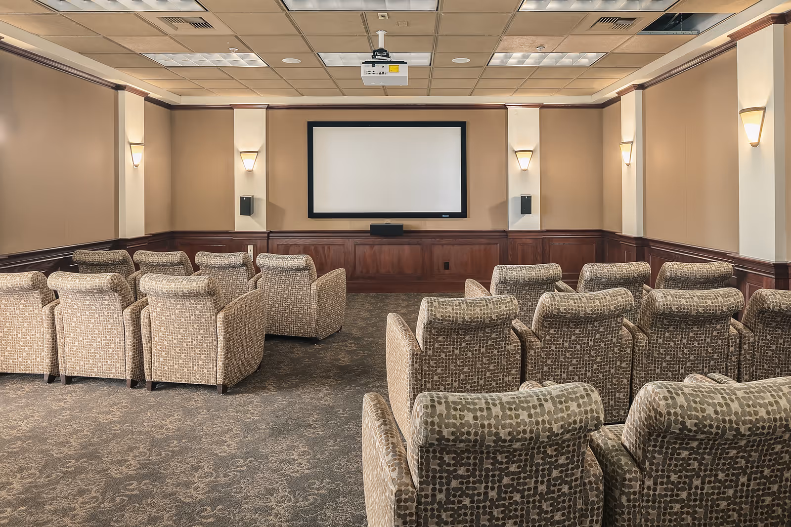Small screening room with rows of patterned upholstered chairs facing a blank projection screen and a ceiling-mounted projector.