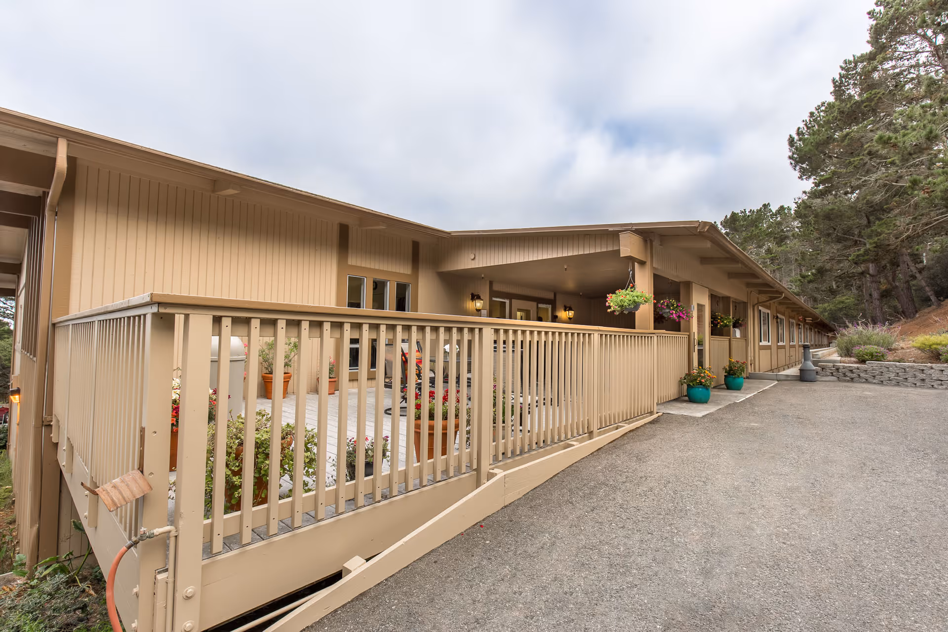 Exterior view of a single-story beige building with a wooden ramp and railing leading to a covered porch area. The porch has hanging flower pots and potted plants along the walkway. Trees and shrubs surround the building under a cloudy sky.