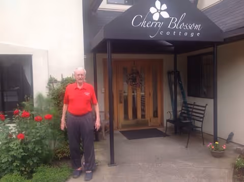 An older man stands in front of the entrance to Cherry Blossom Cottage beneath a black awning, with red flowers and a bench nearby.