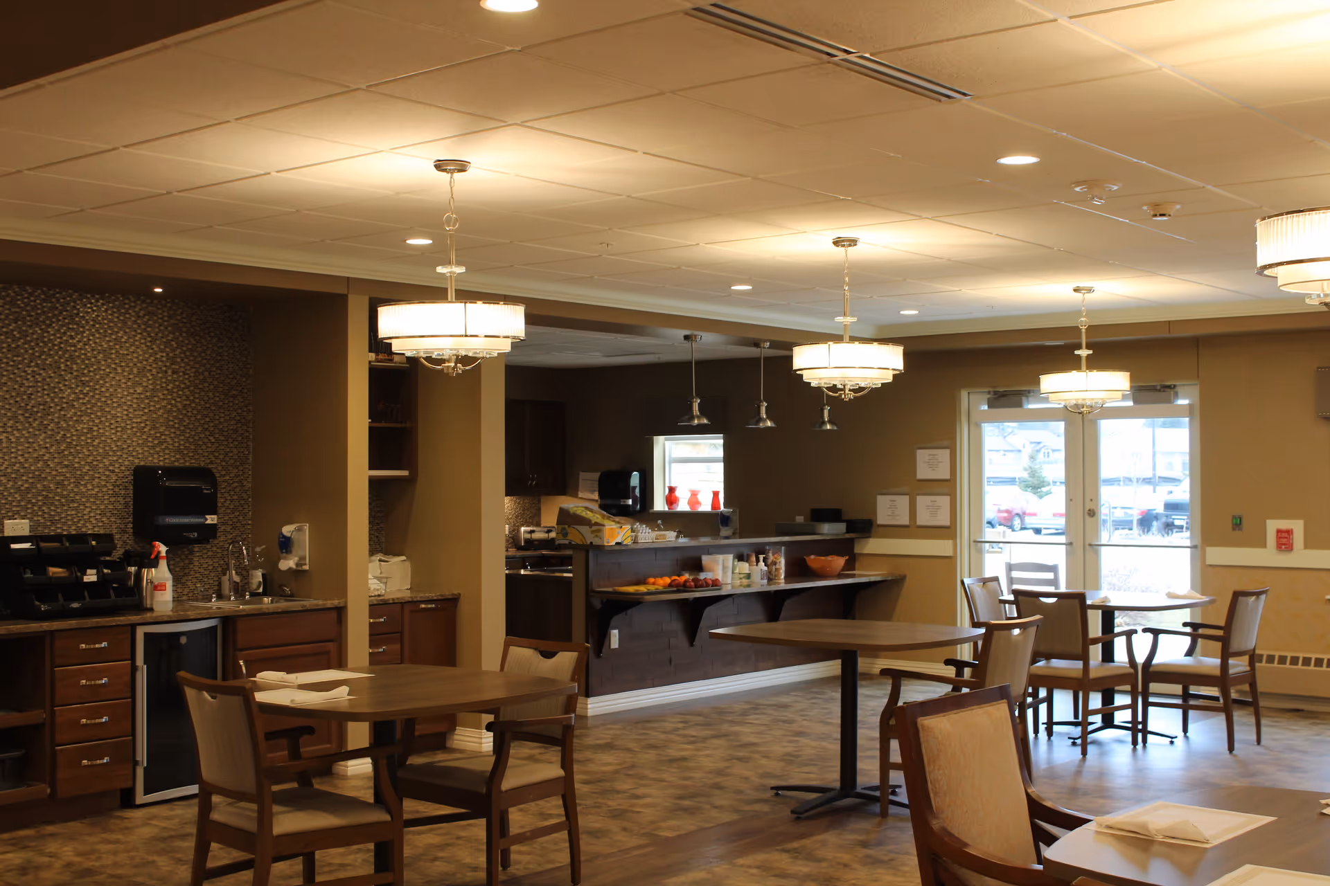 A well-lit dining area in a senior living facility with several wooden tables and chairs. The room features a small kitchen or serving area with cabinets, a sink, and a counter with fruit and other items. There are modern hanging light fixtures and double glass doors leading outside, with a view of parked cars.