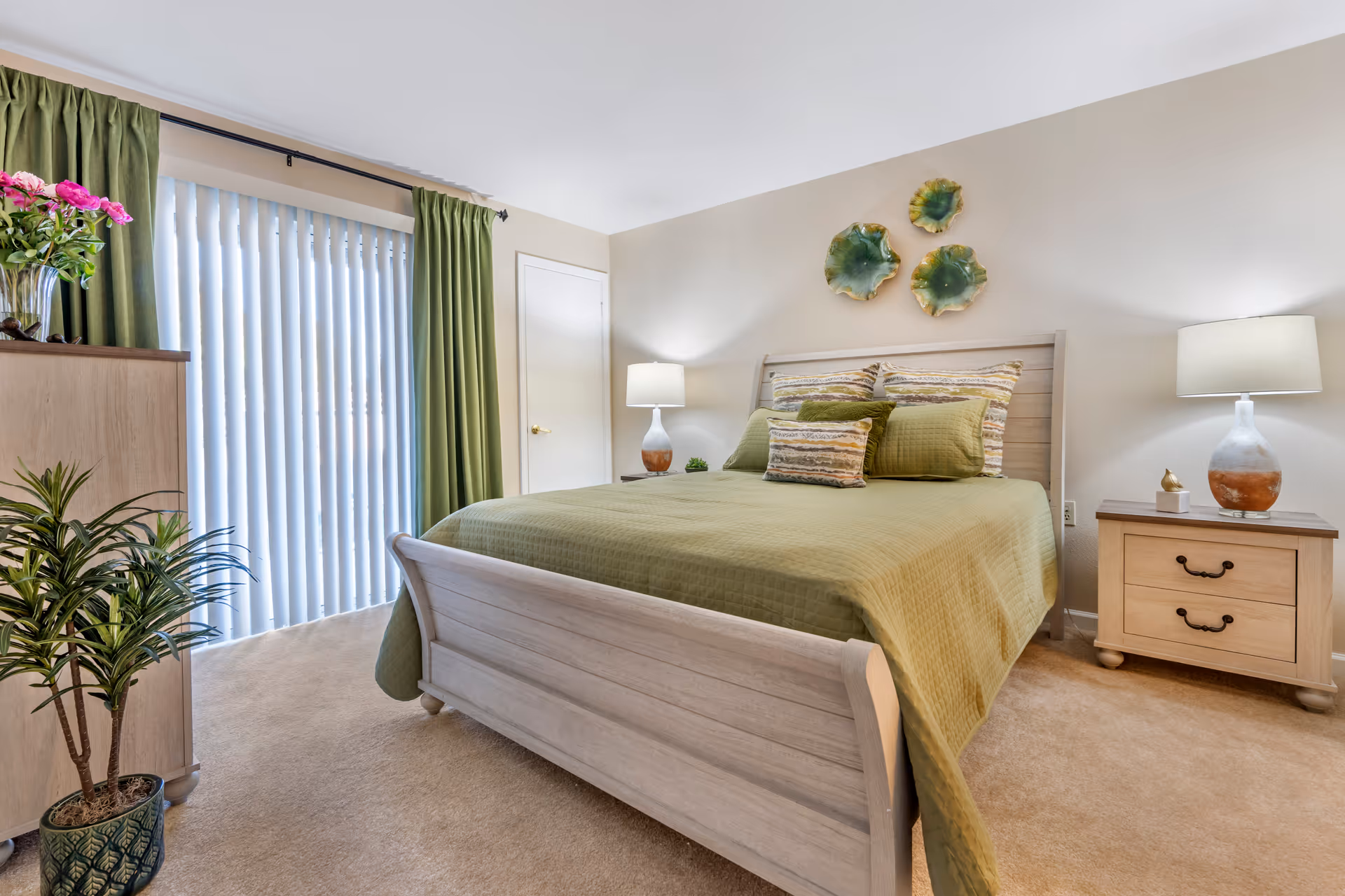 A bedroom with a wooden bed frame and green bedding, including pillows with striped patterns. There are two wooden nightstands on either side of the bed, each with a lamp. Green curtains cover a sliding glass door with vertical blinds. A dresser with a vase of pink flowers and a potted plant are also visible. The walls are light-colored with three green decorative wall hangings above the bed.