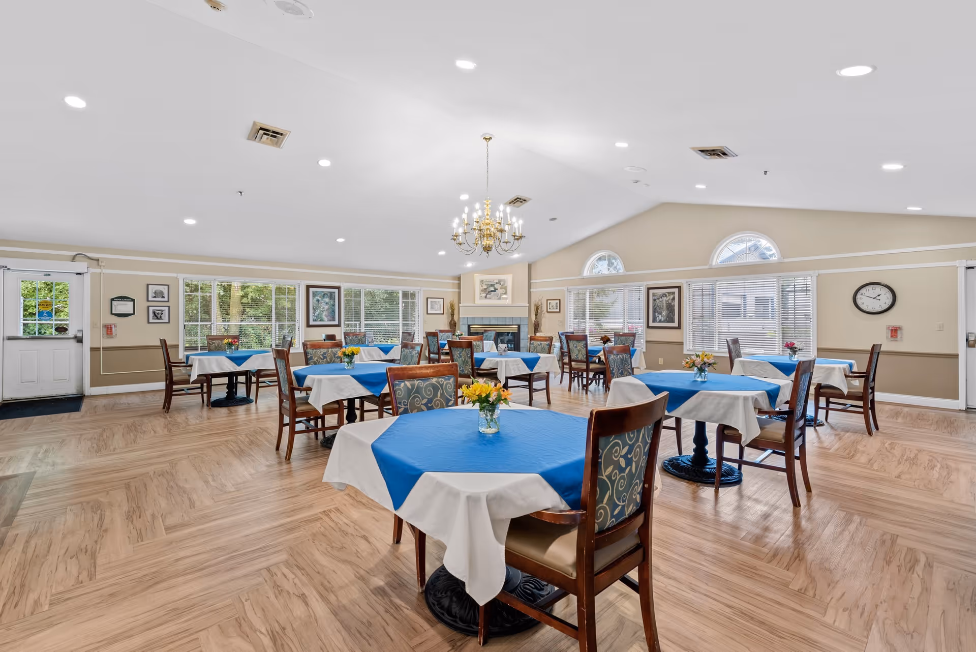A spacious dining room with multiple tables covered with white tablecloths and blue overlays, each adorned with a small vase of flowers. The room features wooden chairs with patterned upholstery, large windows with blinds allowing natural light, a chandelier hanging from the ceiling, and a fireplace at the far end. The floor has a light wood pattern, and the walls are painted beige with white trim.