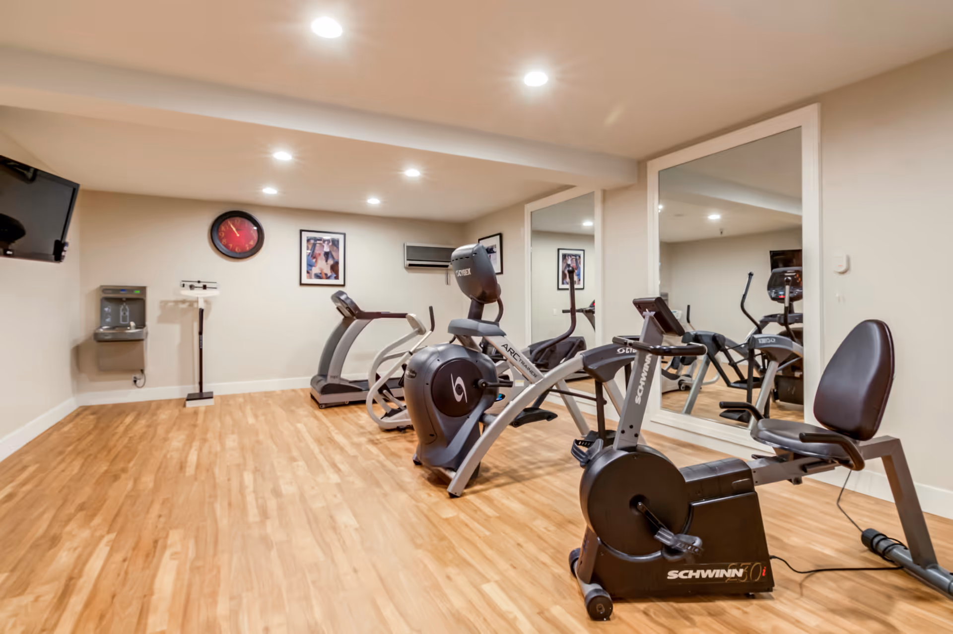 A small fitness room with wooden flooring, containing exercise equipment including a recumbent bike, an arc trainer, and a treadmill. The room has beige walls, a large mirror on one side, a wall-mounted TV, a water fountain, a scale, and framed pictures on the walls. Ceiling lights illuminate the space.