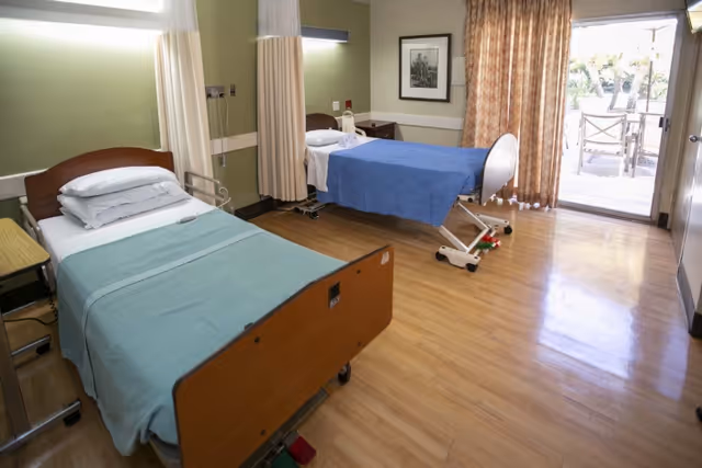 A room with two hospital-style beds, one with a blue blanket and the other with a green blanket, both with pillows. The room has wooden flooring, a framed picture on the wall, a curtain partially drawn to reveal a glass door leading to an outdoor patio with chairs and a table.