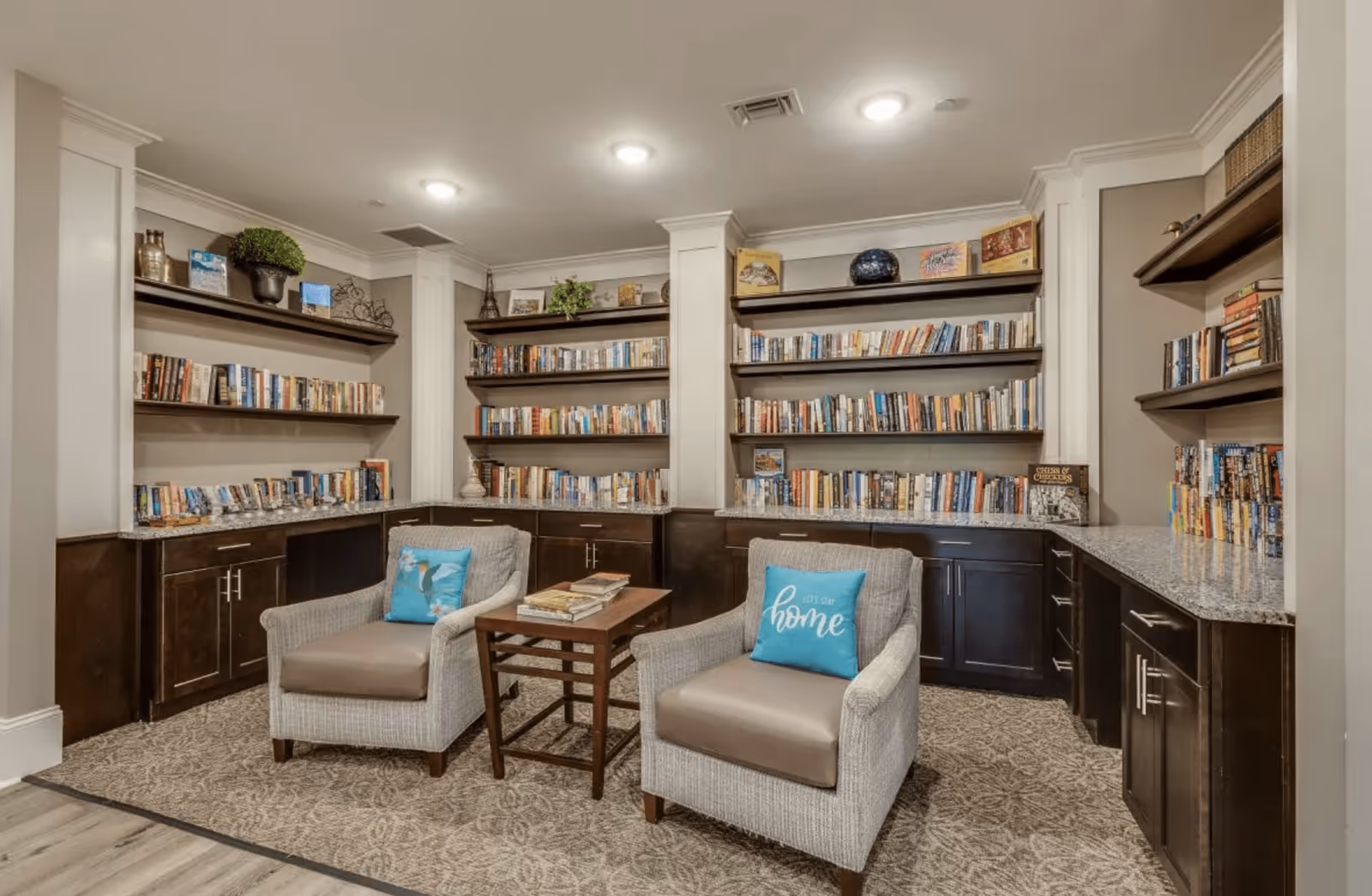 A cozy library or reading nook in an assisted living facility featuring built-in dark wood bookshelves filled with books, two cushioned armchairs with blue pillows, and a small wooden table between the chairs. The room has soft lighting and a patterned carpet.