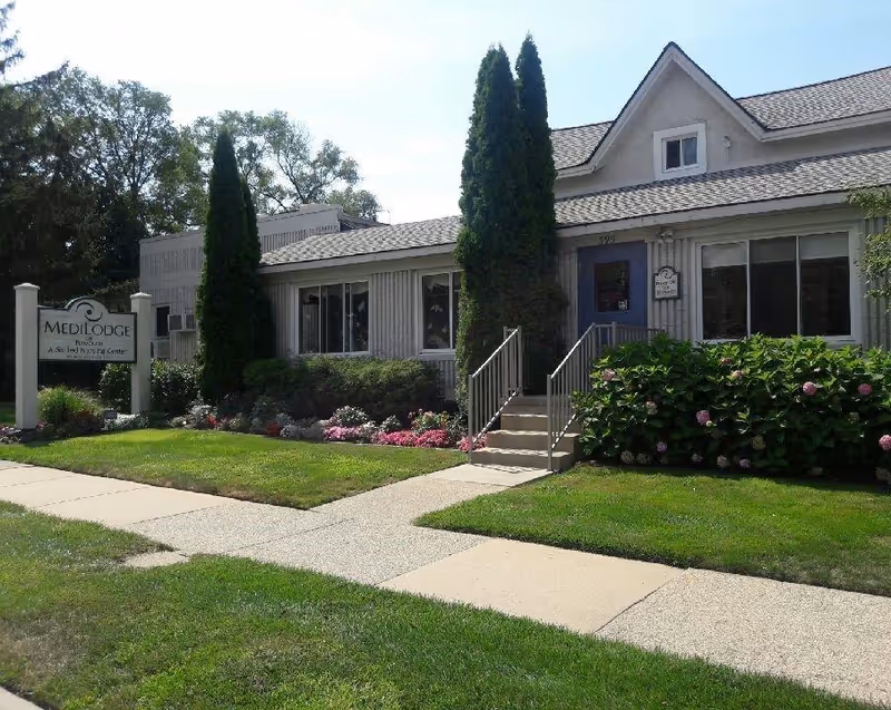 Exterior view of Medilodge of Plymouth, a single-story building with a gabled roof, surrounded by green grass, bushes, and flowers. There is a sidewalk leading to the entrance with stairs and handrails. A sign near the sidewalk reads 'Medilodge Plymouth A Skilled Nursing Center.'