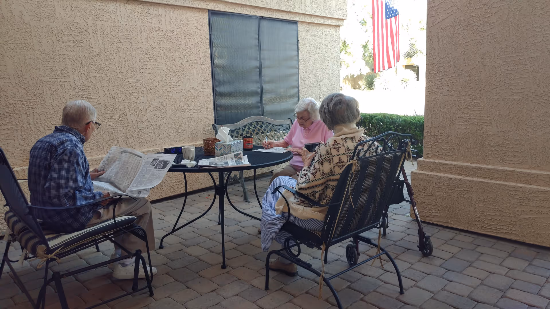 Three elderly residents sit around a round table on a paved covered patio, reading and writing while an American flag hangs nearby.
