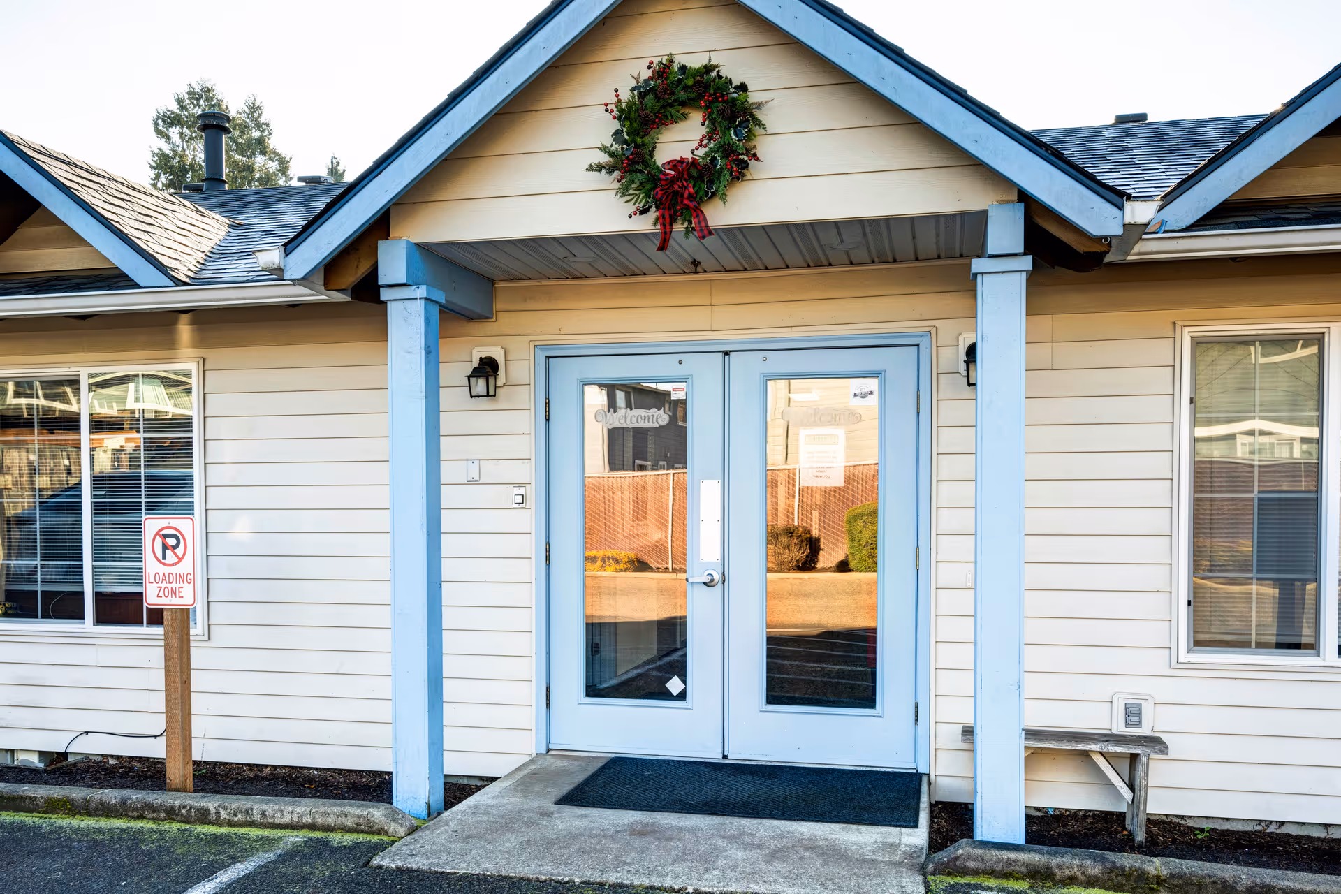 Entrance of a building with double glass doors framed in light blue, a holiday wreath hanging above the doors, and a 'No Parking Loading Zone' sign on a post to the left. The building exterior is light beige with blue trim and two windows on either side of the entrance.