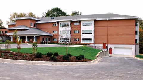 Front exterior of a multi-story brick senior living building with a driveway, lawn, and main entrance.