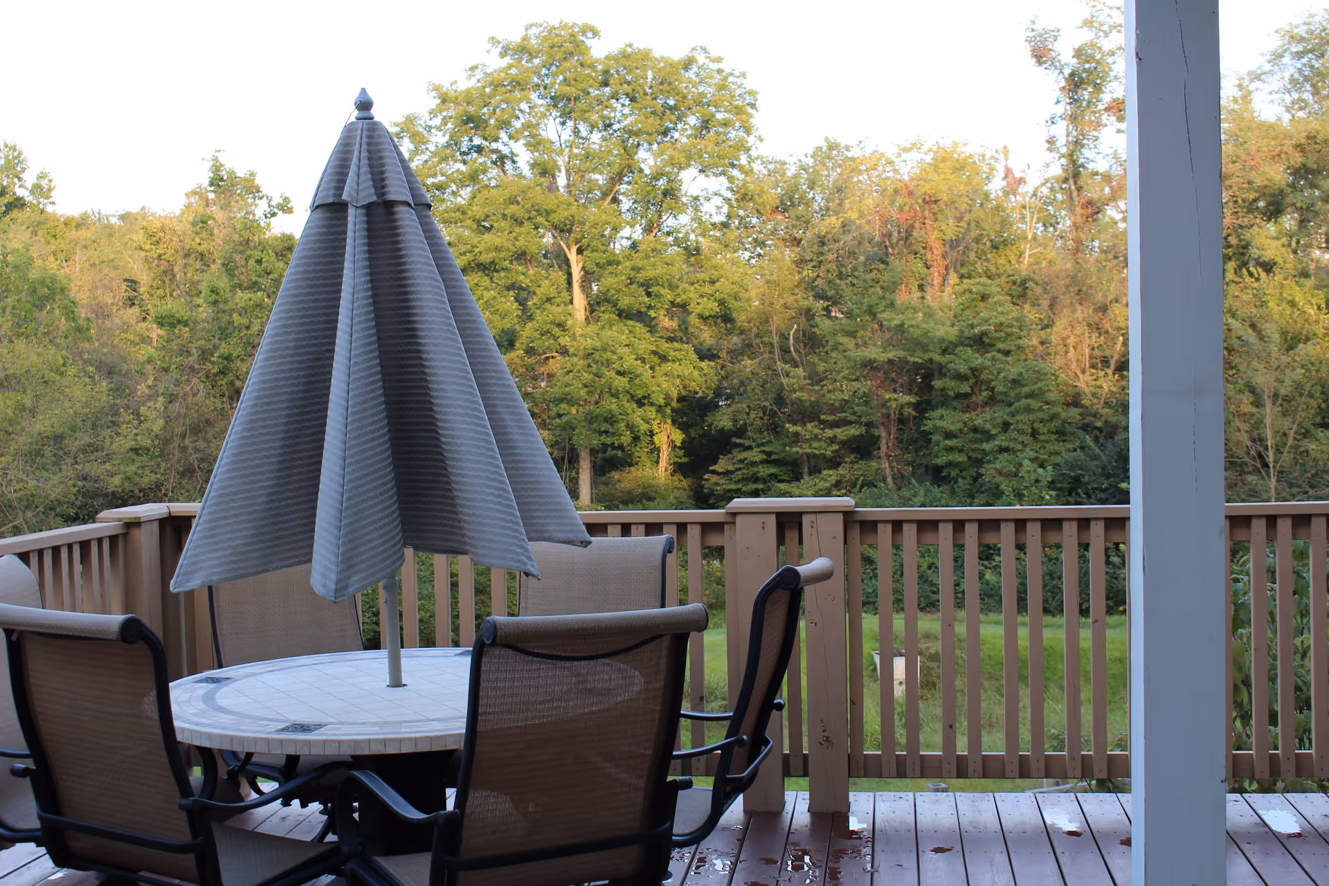A wooden deck with a patio table, chairs, and a closed umbrella overlooking a wooded yard.