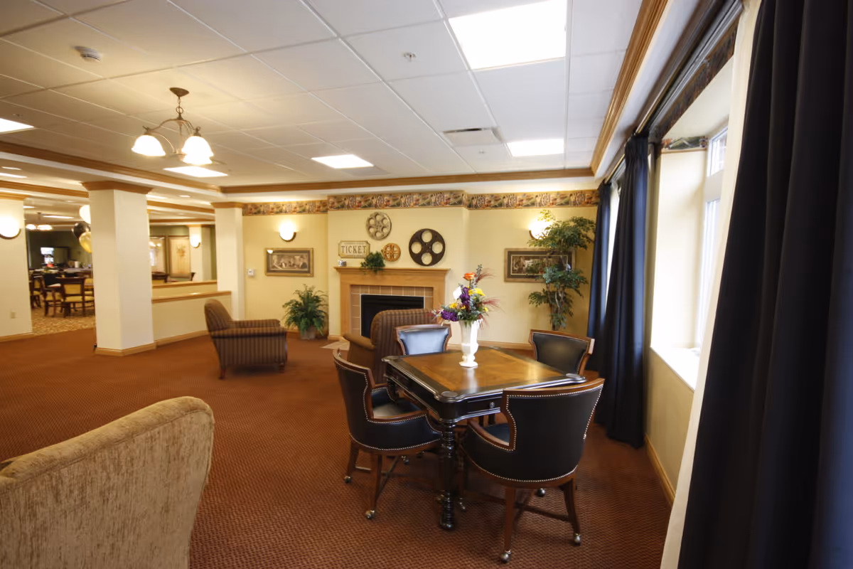 A cozy common area in a senior living facility featuring a square wooden table with four leather chairs, a fireplace decorated with wall art and plants, and large windows with dark curtains allowing natural light to enter. The room has warm lighting, carpeted floors, and additional seating with armchairs in the background.