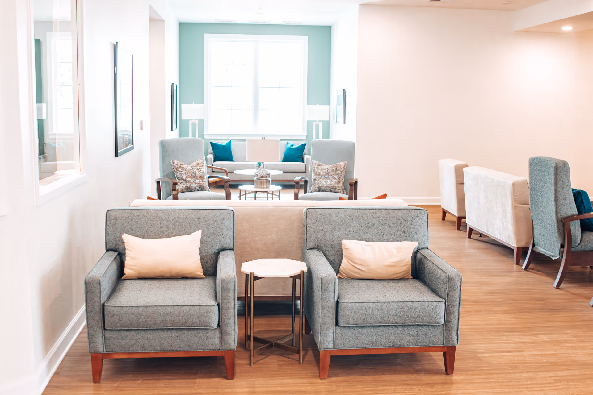 A bright and spacious living room area with two gray armchairs with light pink pillows in the foreground, a small hexagonal side table between them, and a beige sofa behind. In the background, there are two more armchairs with patterned pillows, a sofa with teal pillows, a coffee table, and large windows letting in natural light. The room has wooden flooring and light-colored walls.