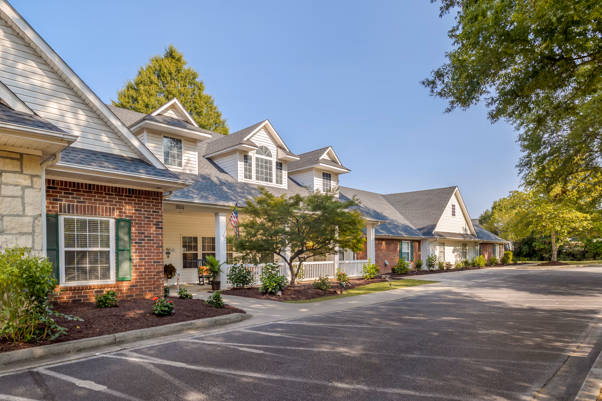 Exterior view of a senior living facility building with brick and siding facade, multiple windows, a porch with white railings, and a tree in front. The parking lot is empty and the sky is clear and blue.