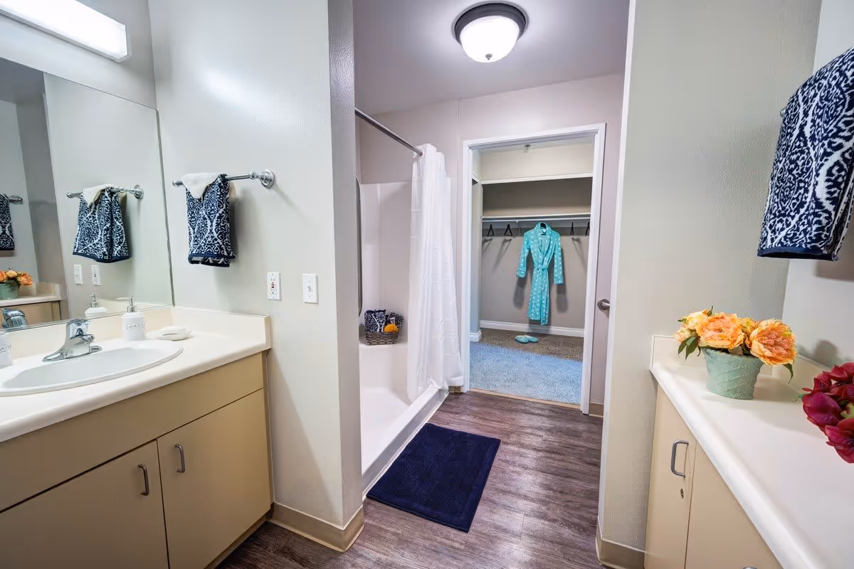 A clean and well-lit bathroom with a sink and countertop on both sides, a large mirror, and a shower with a white curtain. A blue bath mat is placed on the wooden floor in front of the shower. In the background, there is an open doorway leading to a walk-in closet with a teal robe hanging on a hanger and slippers on the carpeted floor.