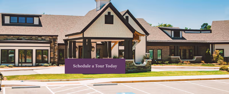 Exterior view of Westminster Memory Care facility showing a large building with a peaked roof, multiple windows, and a covered entrance. The foreground includes a parking lot with marked spaces and a grassy area with landscaping.