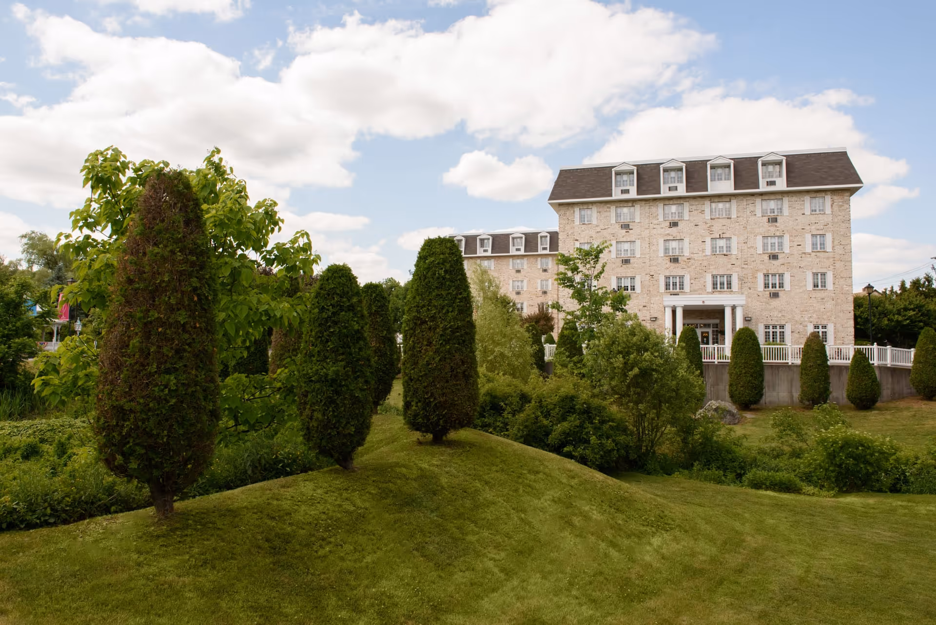 A multi-story beige brick building with white trim, surrounded by well-maintained green lawns and neatly trimmed tall bushes under a partly cloudy sky.