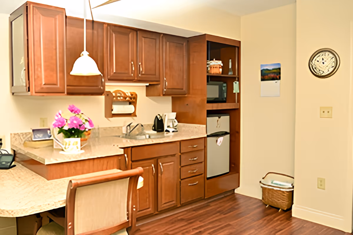 A small kitchen area with wooden cabinets, a countertop with a sink, a coffee maker, a toaster, and a small refrigerator. There is a pendant light hanging above the counter, a chair with a cushioned back, a basket on the floor, a wall clock, and a calendar on the wall.