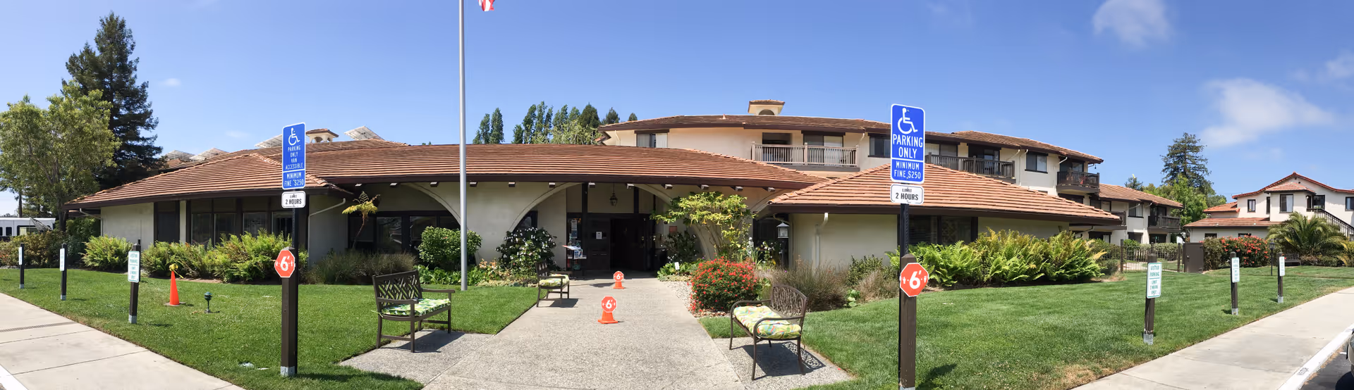 Front exterior view of La Posada Retirement Community (Independent Living) showing a single-story building with a tiled roof, benches along the walkway, green lawns, and handicap parking signs under a clear blue sky.