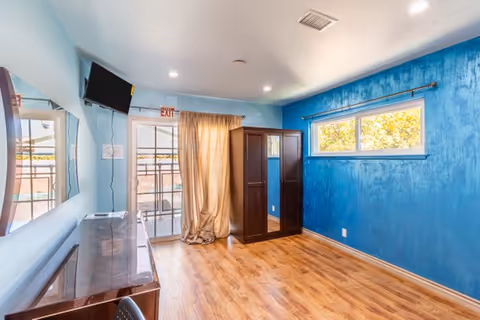 Empty furnished room with wood floors, a blue accent wall, wardrobe, desk, wall-mounted TV and sliding glass door with curtains.