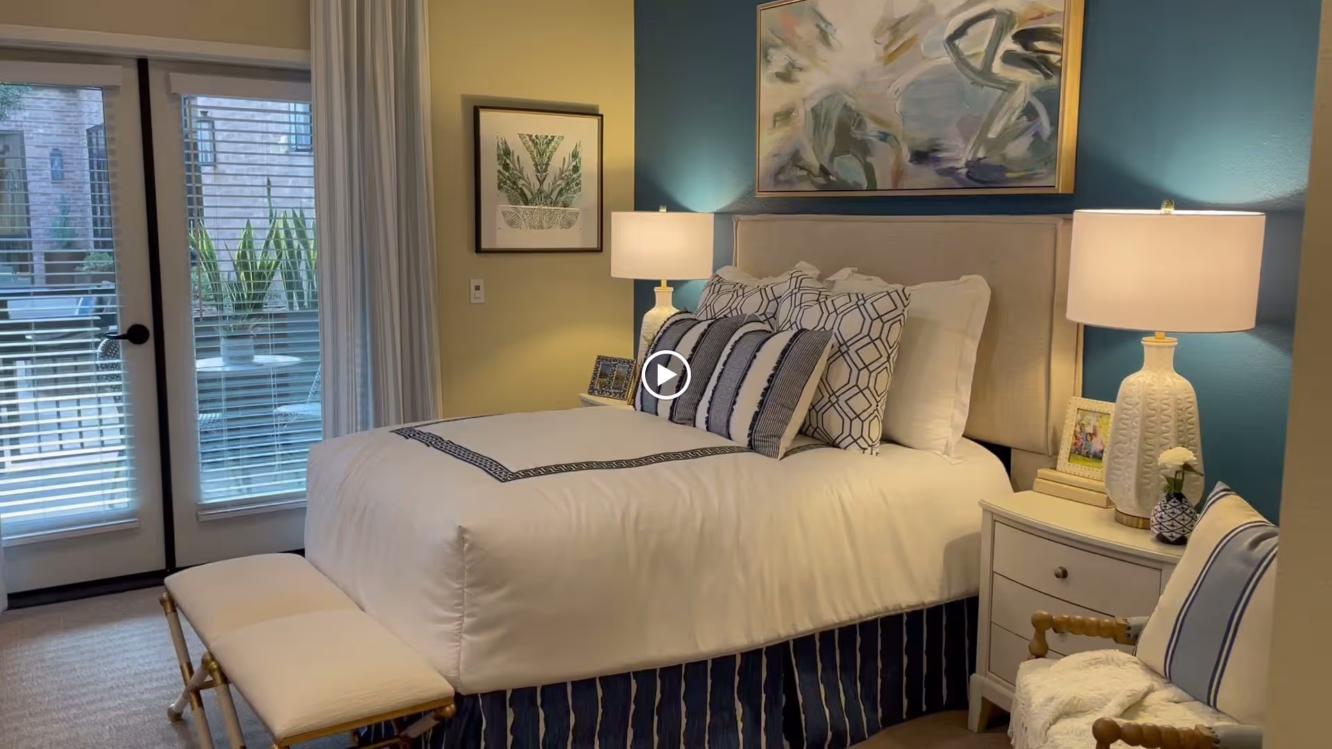 A neatly made bed with white and blue bedding in a bedroom. The bed has multiple decorative pillows and a padded headboard. There are two bedside tables, each with a lamp and decorative items. A framed abstract painting hangs above the bed, and a framed botanical print is on the adjacent wall. French doors with blinds lead to an outdoor patio with plants visible outside. A cushioned bench is at the foot of the bed, and a chair with a striped pillow and a throw blanket is in the corner.