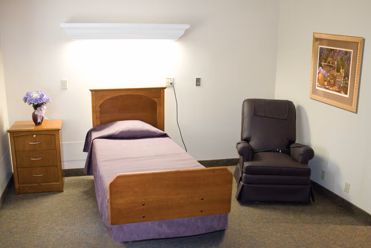 A simple bedroom in an assisted living facility featuring a single wooden bed with a purple bedspread, a wooden nightstand with a vase of purple flowers, a dark brown recliner chair, and a framed floral painting on the wall.