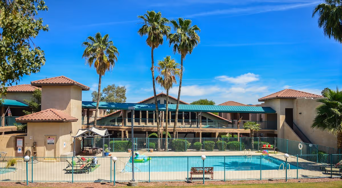 Outdoor swimming pool area at a senior living facility with lounge chairs, palm trees, and a two-story building with a green roof and beige walls in the background under a clear blue sky.