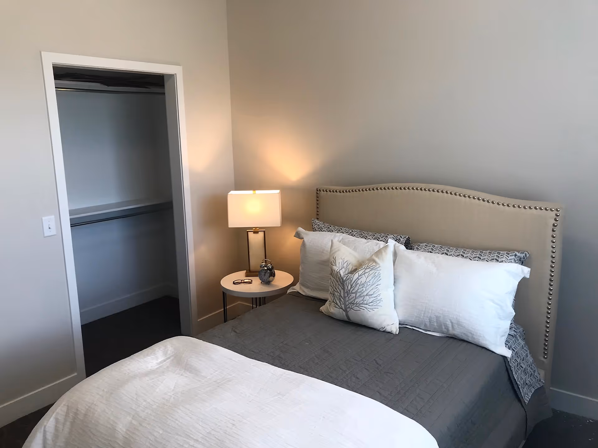 A bedroom with a neatly made bed featuring a beige upholstered headboard with nailhead trim, gray and white bedding, and decorative pillows. A small round bedside table with a lamp, glasses, and a small decorative item is next to the bed. An open closet with a hanging rod and shelf is visible on the left side of the room.