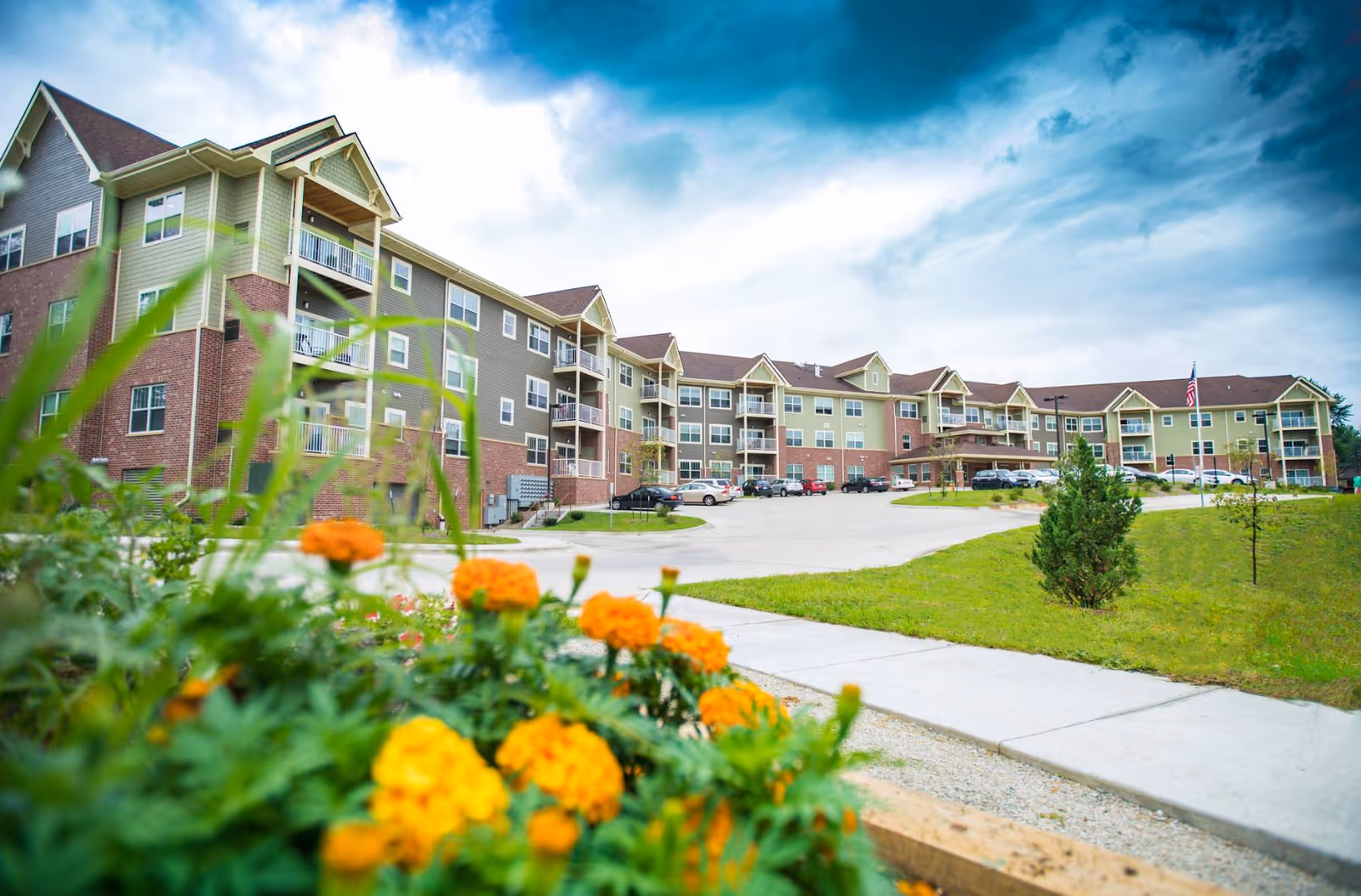 Exterior view of a multi-story senior living facility building with balconies, a parking lot with cars, a sidewalk, green grass, and orange flowers in the foreground under a partly cloudy sky.