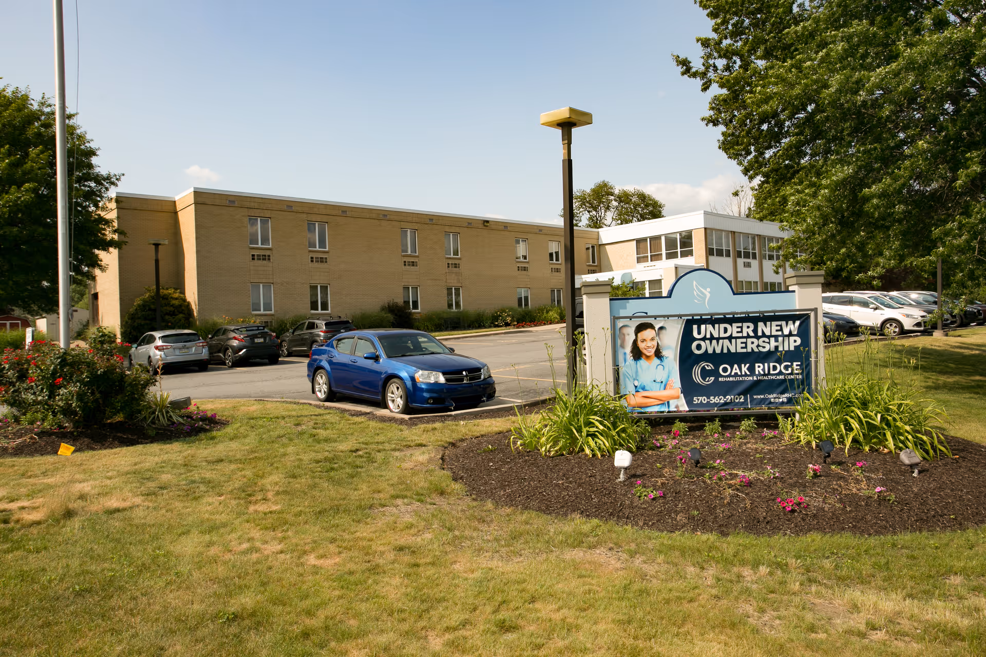Exterior view of Oak Ridge Rehabilitation and Healthcare Center showing a two-story building with multiple windows, a parking lot with several cars, a landscaped area with flowers and grass, and a sign indicating the facility is under new ownership.