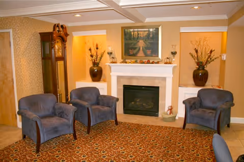 A cozy living room area with three blue upholstered armchairs arranged around a fireplace. The fireplace has a white mantel decorated with small pumpkins and a framed painting above it. On either side of the fireplace are built-in alcoves with large vases holding decorative dried plants. A tall grandfather clock stands to the left of the fireplace. The room has warm beige walls and a patterned carpet.