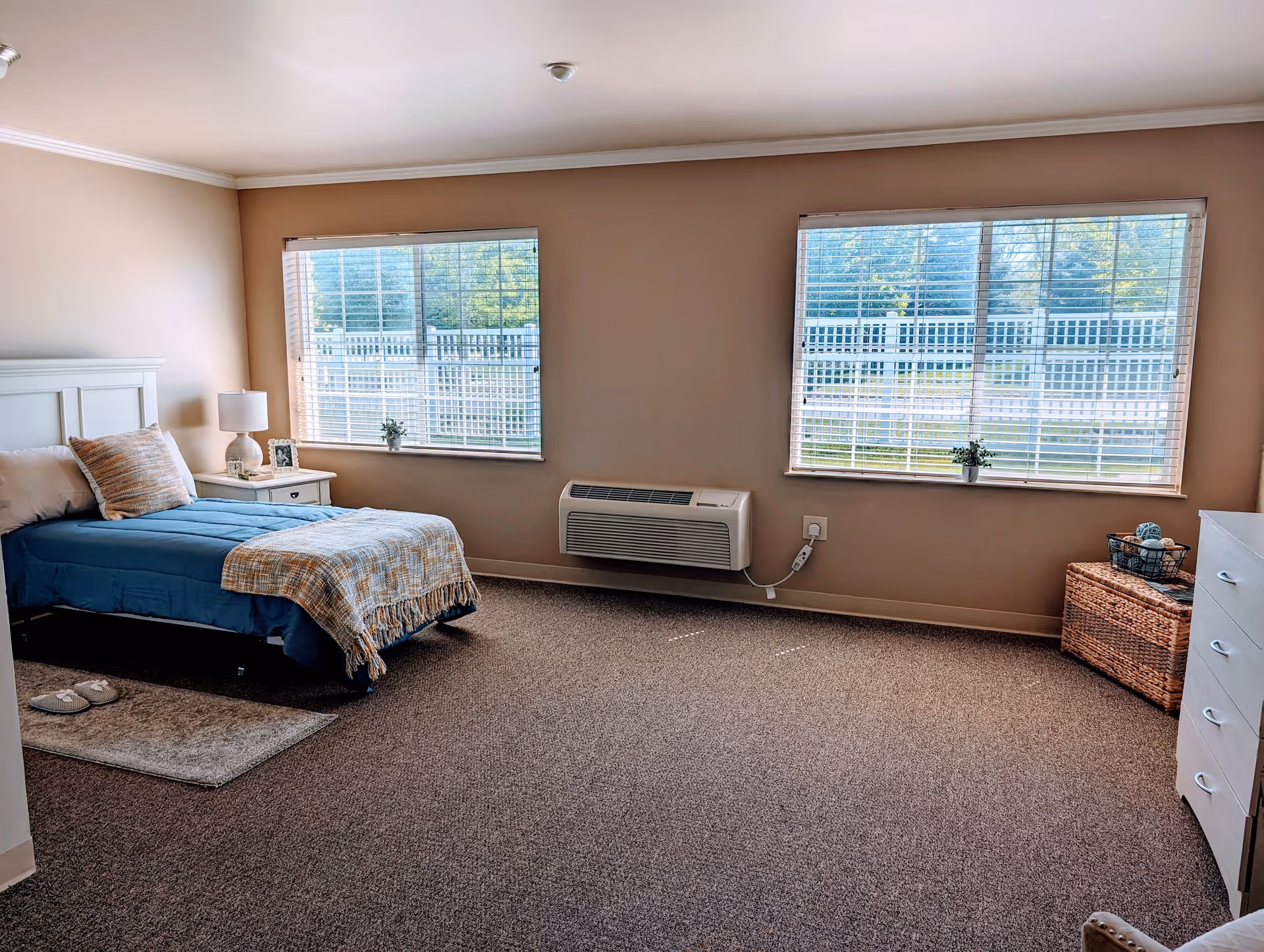 A neatly arranged bedroom with a single bed covered in blue bedding and a beige throw blanket. There is a white nightstand with a lamp, a small plant, and a framed photo next to the bed. Two large windows with white blinds let in natural light, showing a view of a white fence and greenery outside. A wicker basket and a white dresser with drawers are also visible in the room, which has beige walls and carpeted flooring.