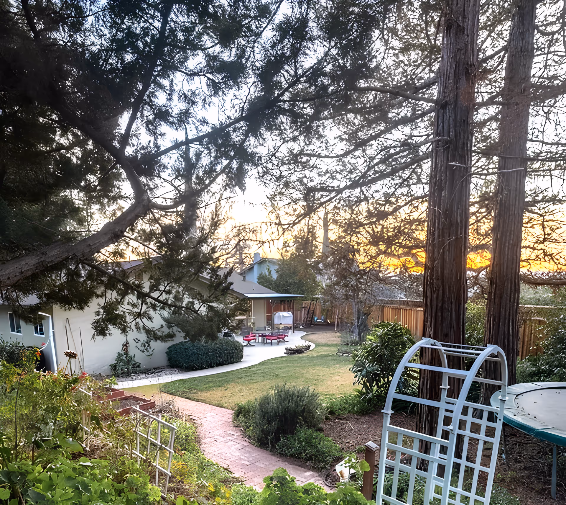 A backyard garden area at sunset with a paved walkway, green lawn, various plants and bushes, two tall trees, a white garden arch, and a trampoline. A single-story house with a patio and outdoor furniture is visible in the background.