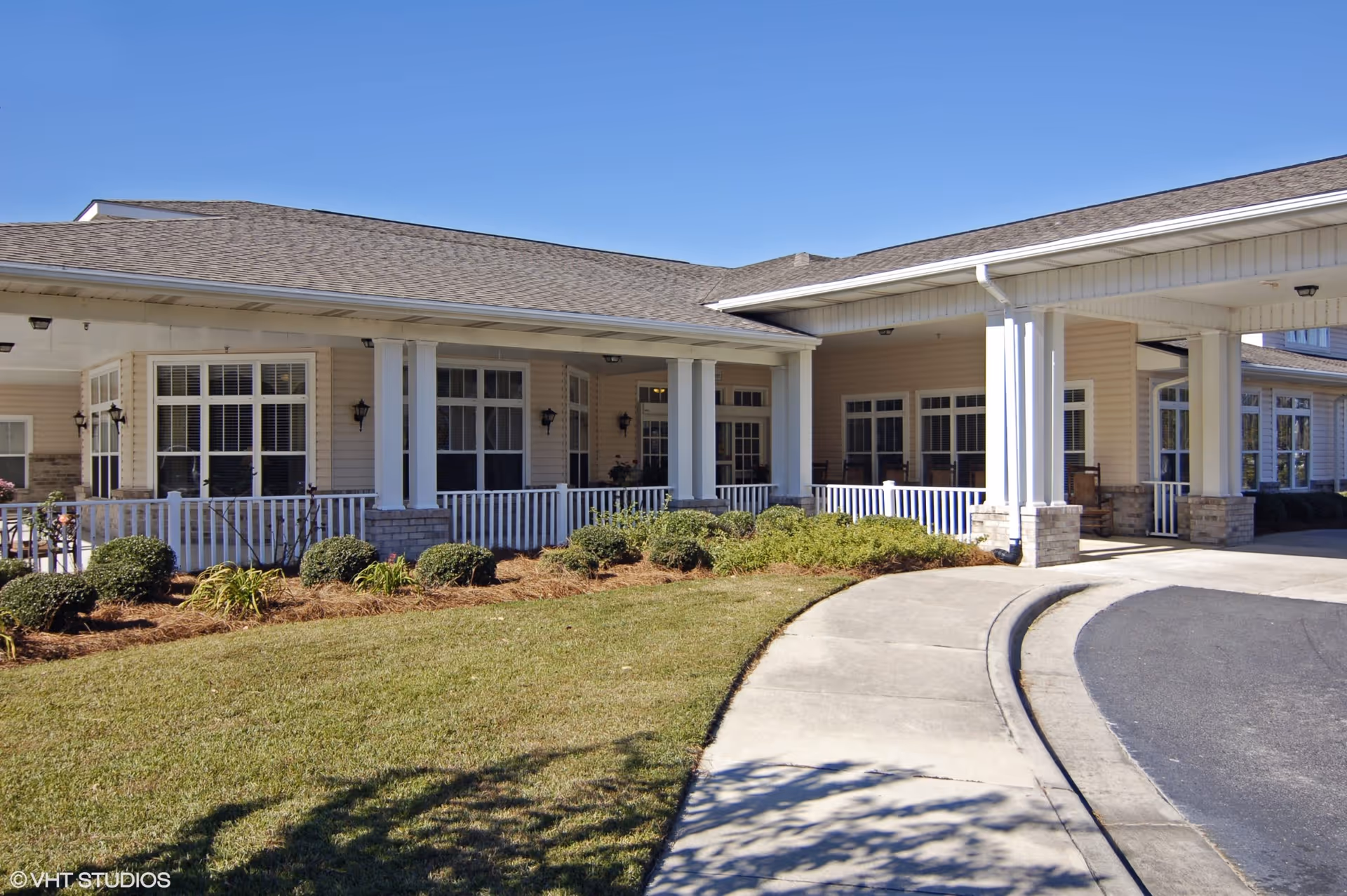 Exterior view of a senior living facility building with beige siding, white columns, and a covered entrance. There are multiple windows with white frames and a landscaped area with bushes and grass in front. The sky is clear and blue.