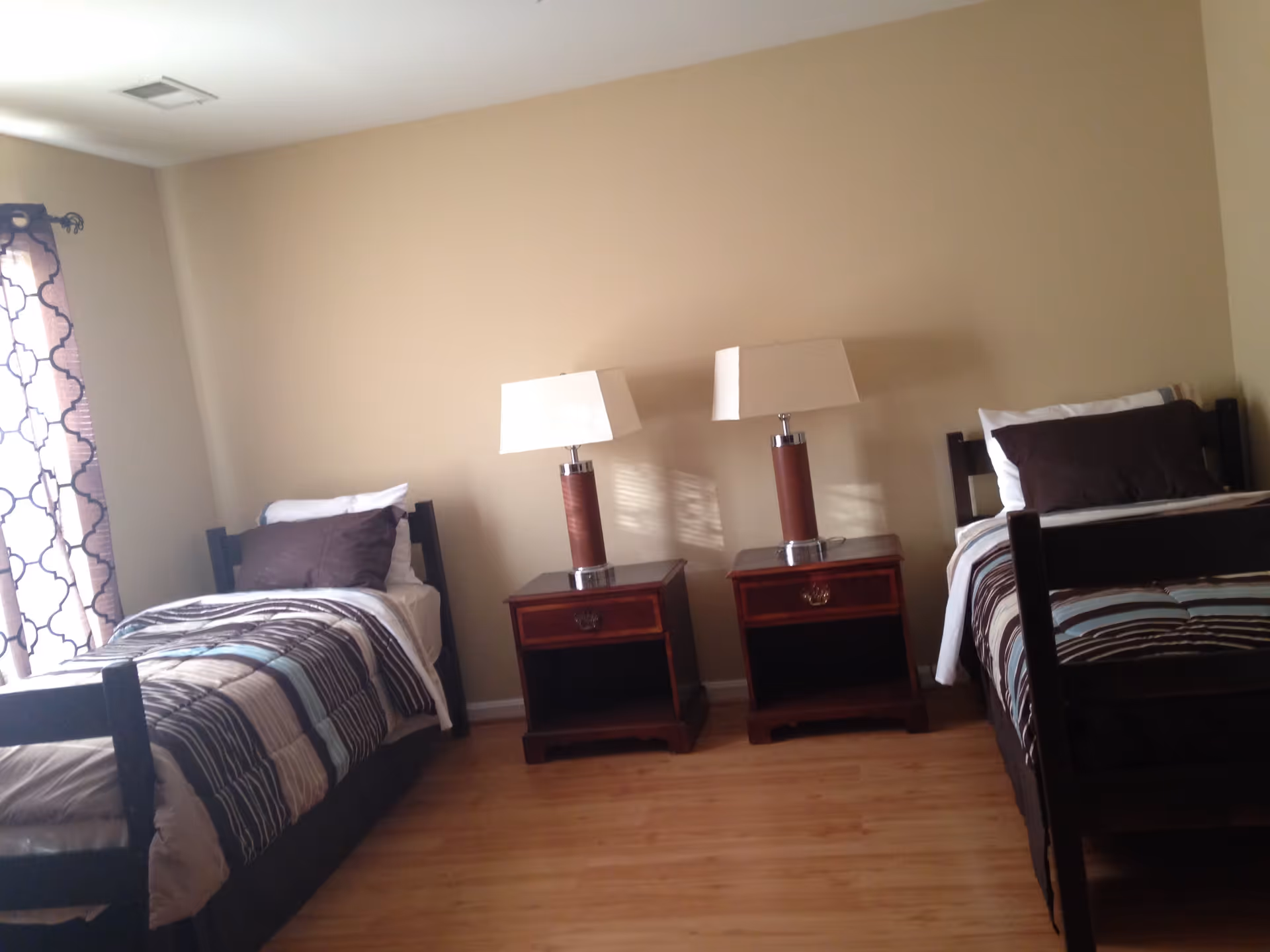 A bedroom with two single beds separated by two wooden nightstands, each with a lamp. The beds have striped bedding and pillows, and there is a window with patterned curtains on the left side. The walls are beige and the floor is wooden.