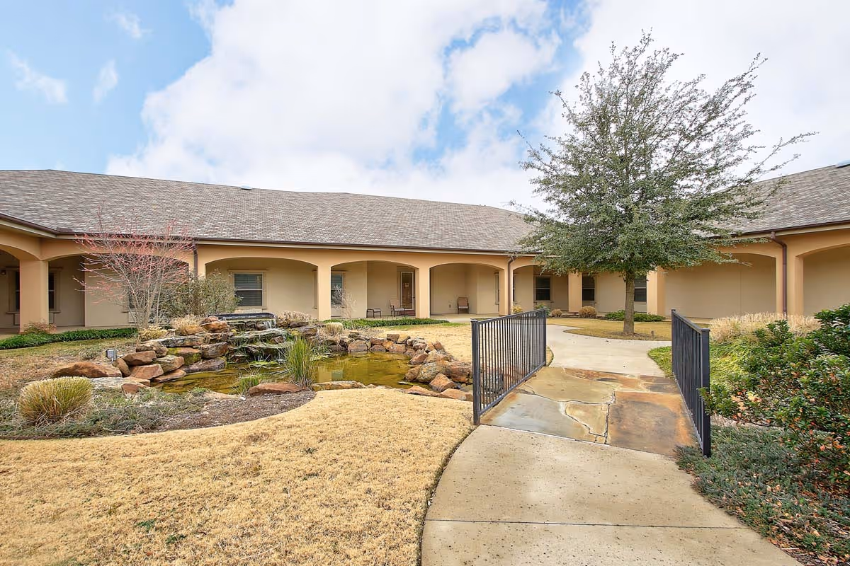 Outdoor courtyard area at The Oaks At Flower Mound featuring a small pond with rocks and plants, a paved walkway with a metal railing, a tree, and a building with a covered walkway and arches in the background under a partly cloudy sky.