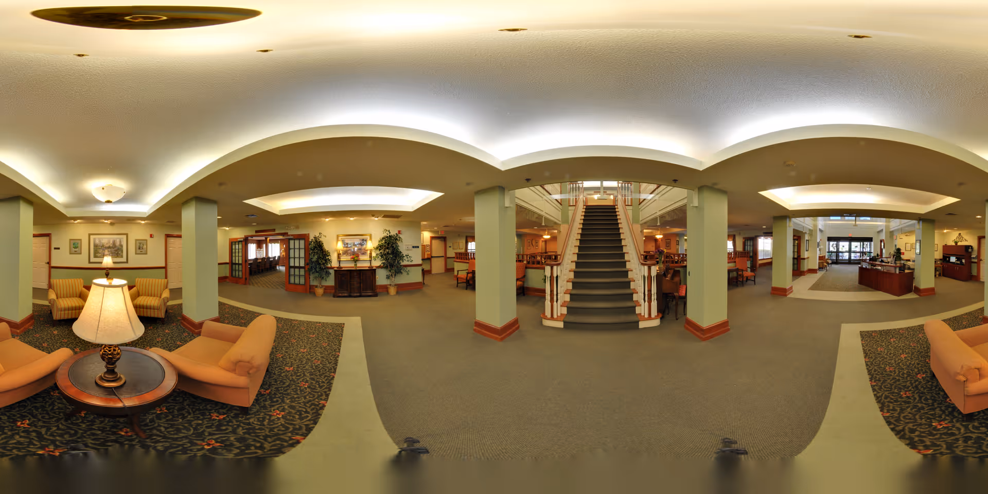 A spacious and well-lit senior living facility common area with a central staircase leading to an upper floor. The room features comfortable seating areas with armchairs and lamps, decorative plants, framed artwork on the walls, and a reception or service desk area to the right. The carpeted floor and soft lighting create a warm and inviting atmosphere.